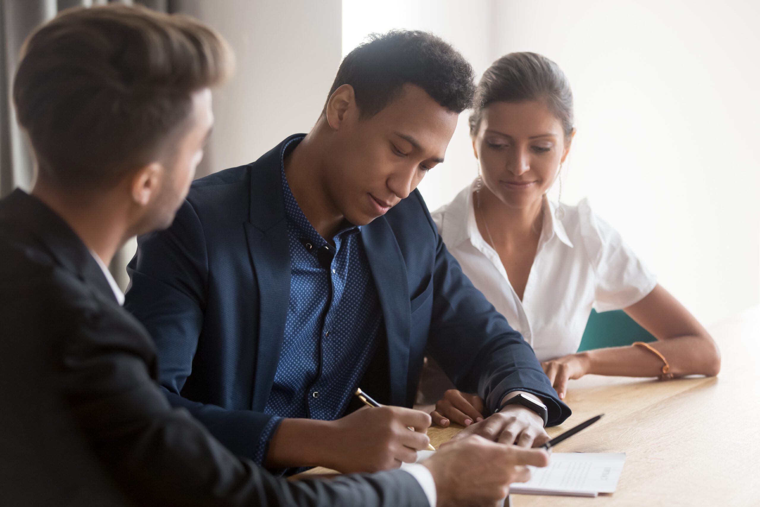 Three people sit at a table, with one man signing a document while the others observe.